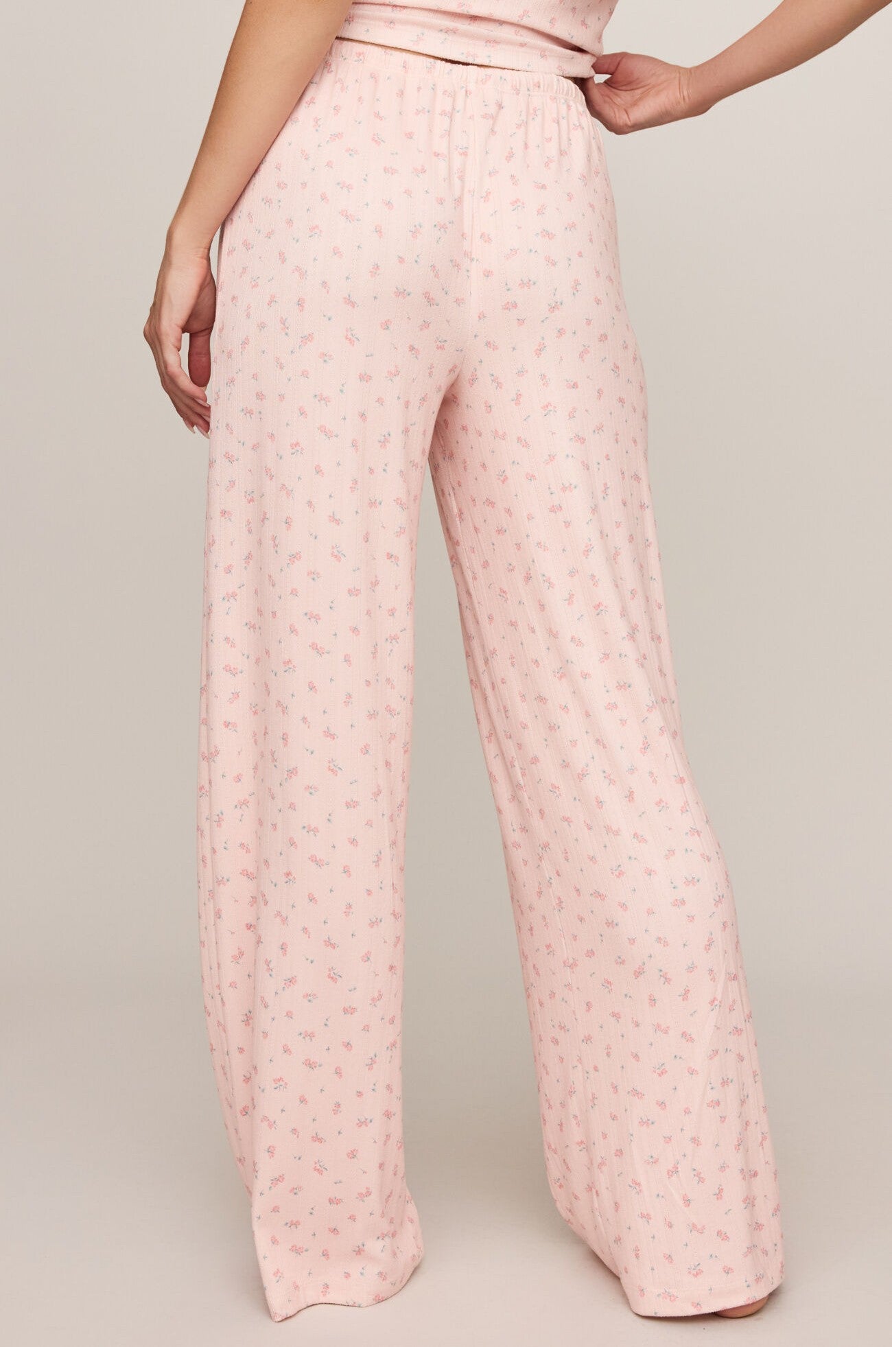 A person stands facing away, wearing Z Supply’s Better Off Ditsy Pant—full-length, high-rise light pink pajama pants with a small floral print and relaxed wide-leg fit. The background is plain and neutral.