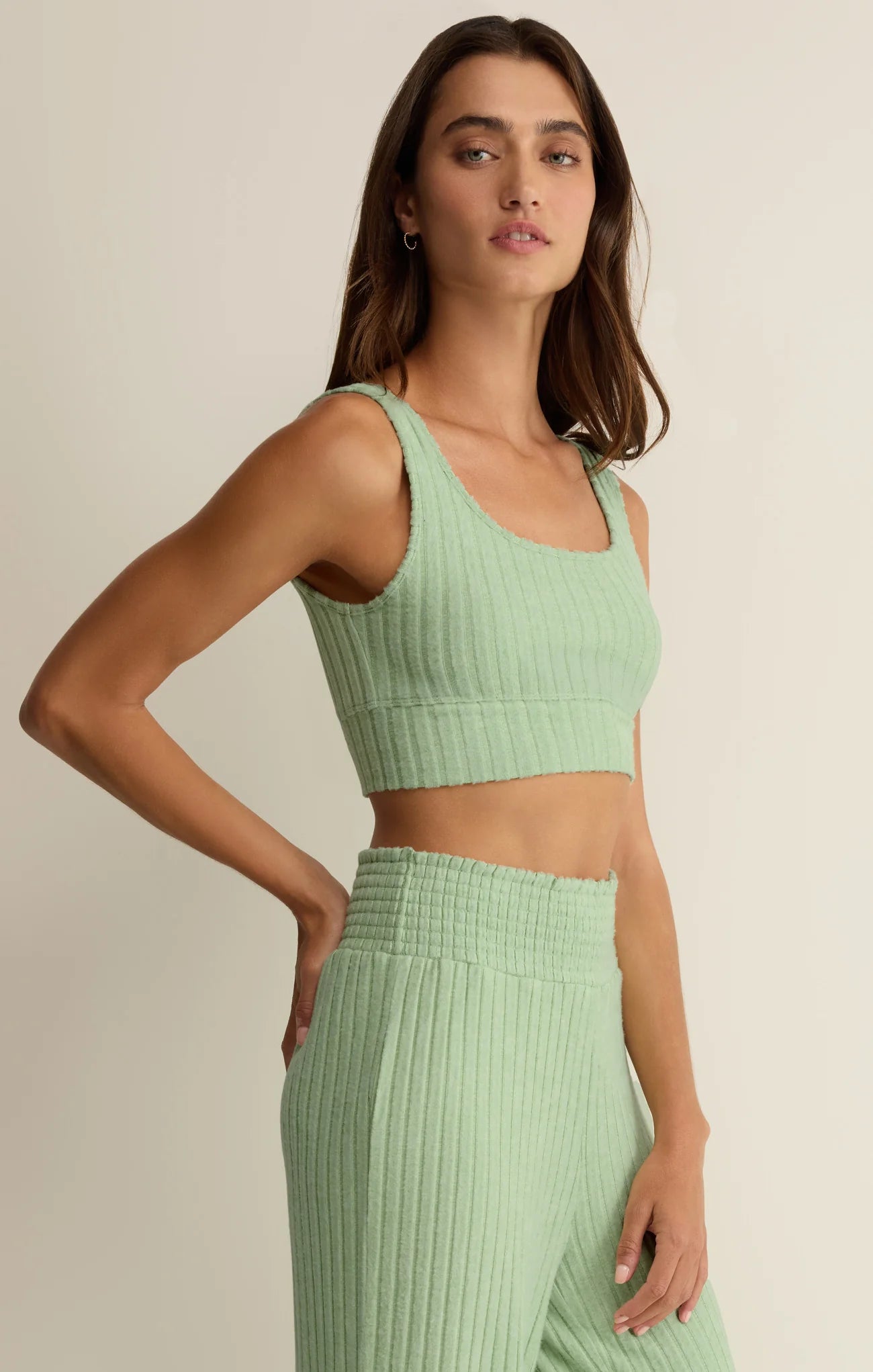A woman wearing the Z Supply Zoe Rib Tank Bra in Sprout, a light green ribbed crop top with light support, stands with one hand on her hip against a plain light background.