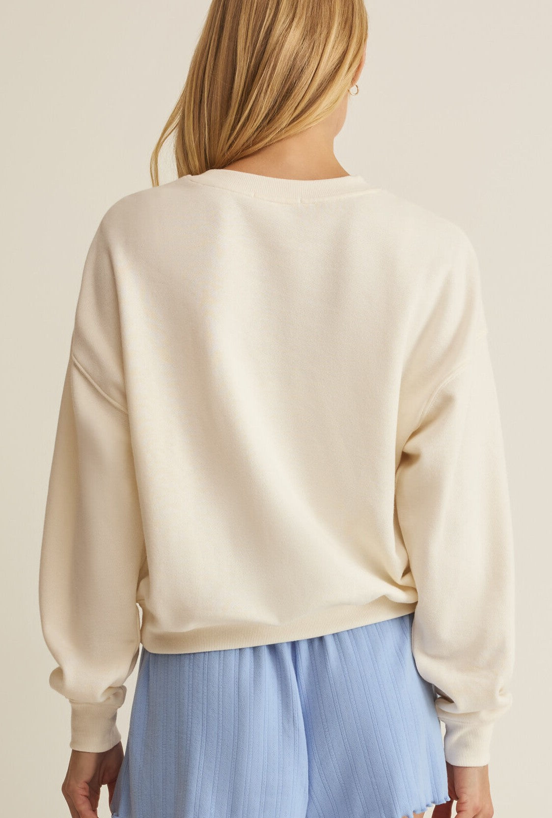 A person with long blonde hair is shown from the back, wearing the Z Supply Mama Sweatshirt and light blue pleated shorts against a plain, neutral background.