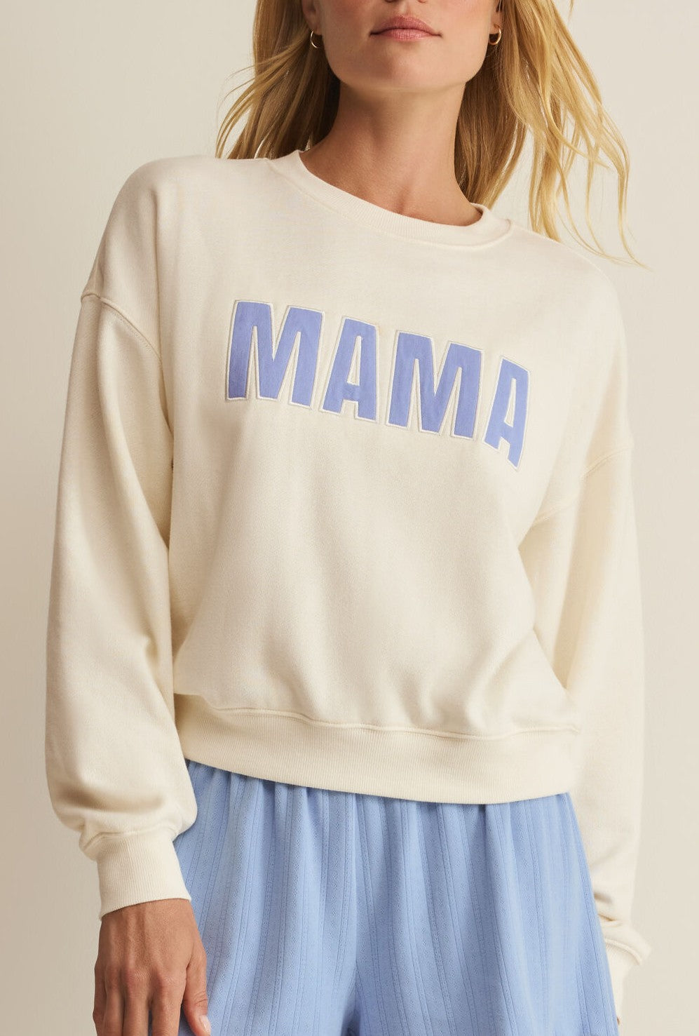 A woman models the Z Supply Mama Sweatshirt, a cream-colored cotton fleece with bold blue "MAMA" lettering, paired with loose light blue pants against a neutral background.