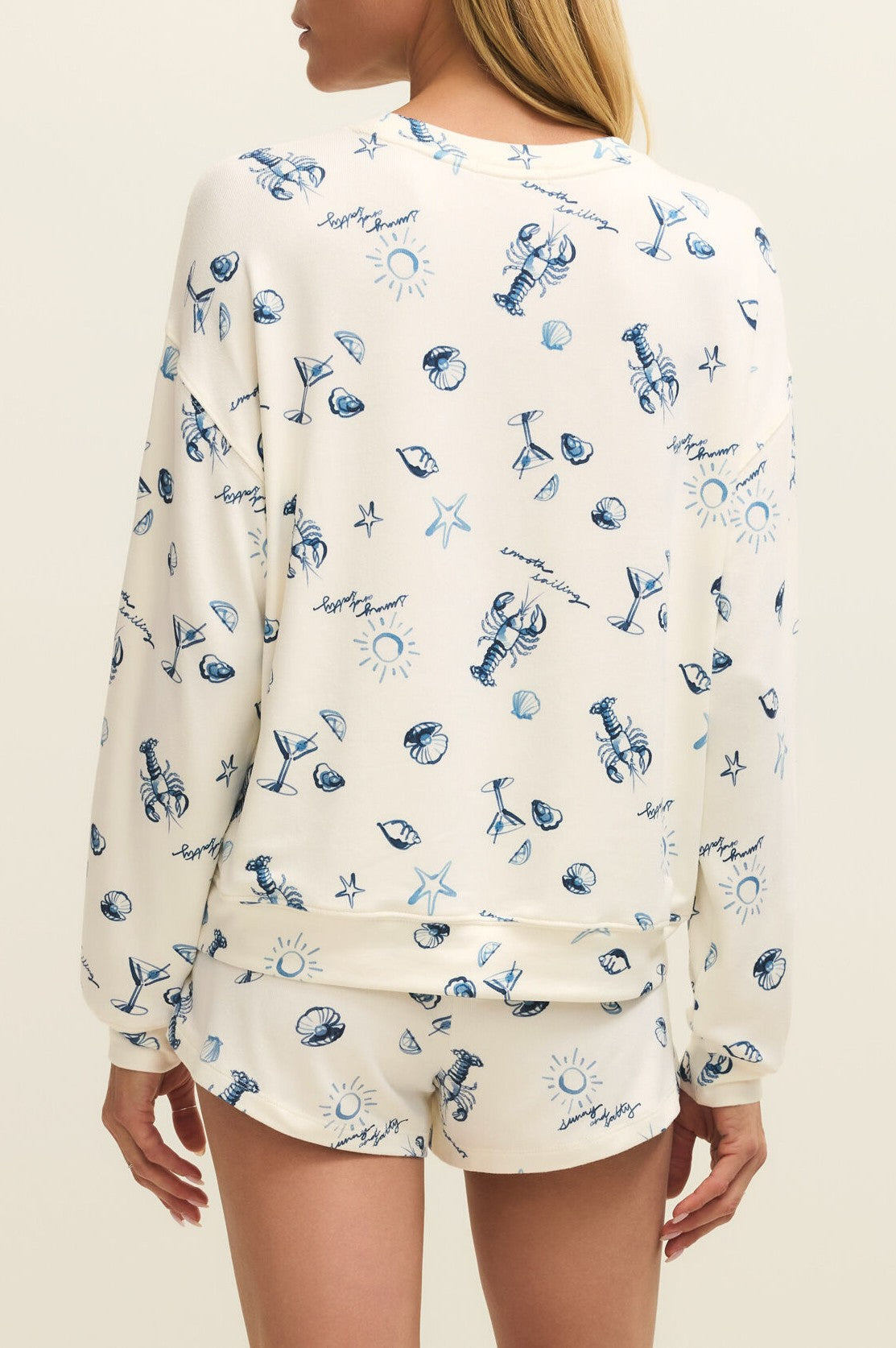 A woman faces away, wearing the Z Supply Lost At Sea Long Sleeve Top—a relaxed fit, cream crewneck pajama top with shorts, made from silky jersey and decorated with blue beach-themed prints like lobsters and shells.