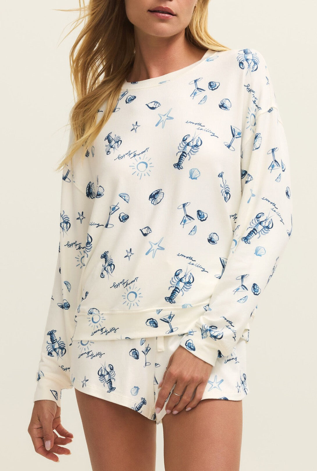 A woman wears the Z Supply Lost At Sea Long Sleeve Top, a relaxed fit cream crewneck with blue nautical prints—lobsters, seashells, and starfish—standing against a plain light background. Her face is partially out of the frame.