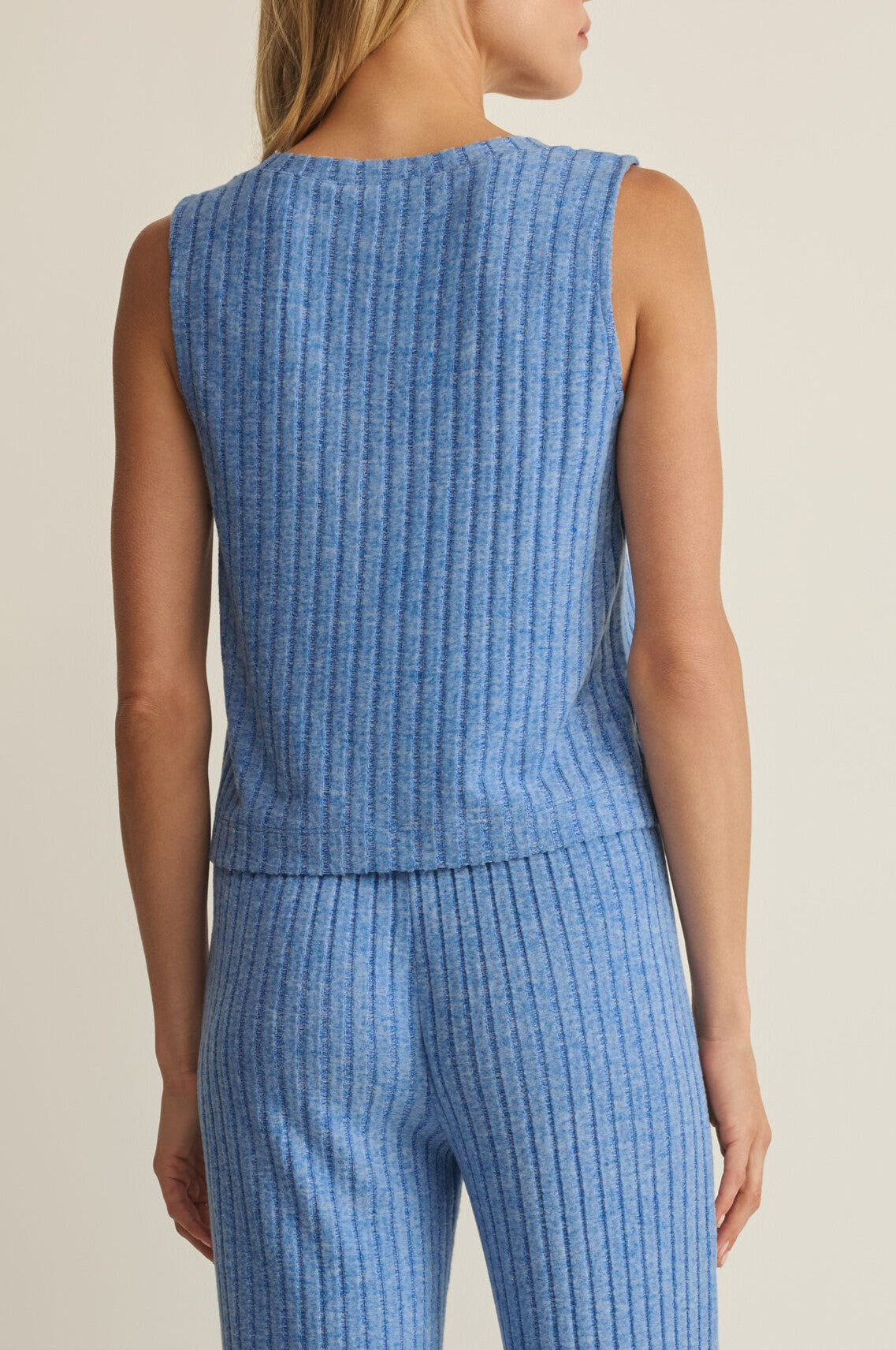 A woman with long blonde hair and light skin is seen from the back wearing the Z Supply Sloane V Neck Rib Tank, a sleeveless relaxed-fit blue knit top, with matching ribbed pants. The background is plain and neutral.