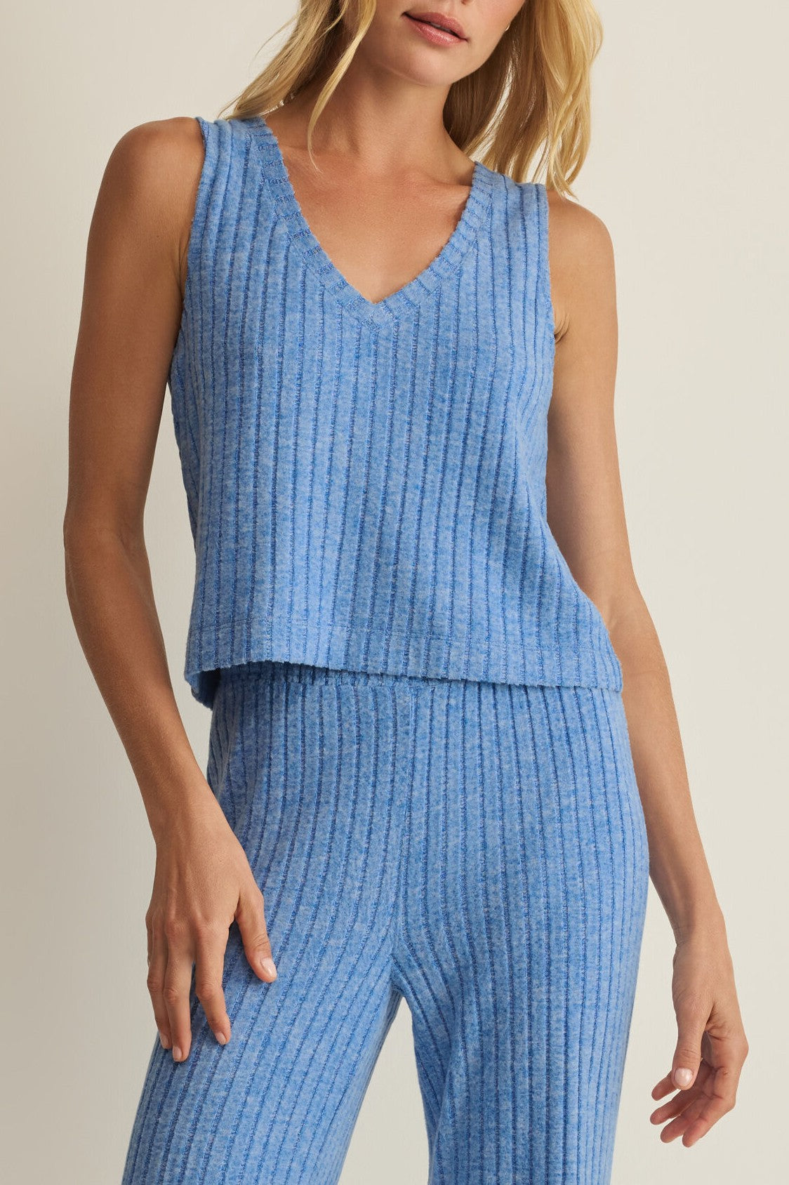 A woman wears the Z Supply Sloane V Neck Rib Tank in pale blue, styled with high-waisted pants. The relaxed, ribbed knit top looks soft and comfortable—ideal loungewear—against a plain, neutral background.