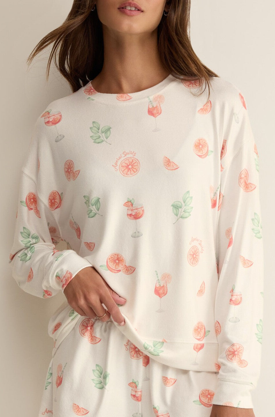 A woman wears the Z Supply Spritz Pj Top in White Multi—relaxed fit, silky jersey featuring grapefruit slices, green leaves, and cocktail glasses—paired with matching pants. She poses against a neutral background to highlight the fun print.