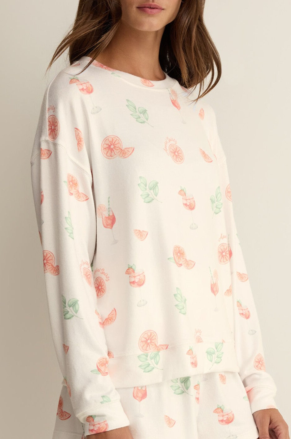 A woman wears the Z Supply Spritz Pj Top, a relaxed fit white long-sleeve shirt with pastel fruit and cocktail glass spritz prints in silky jersey. Her head is partially out of frame against a neutral background.