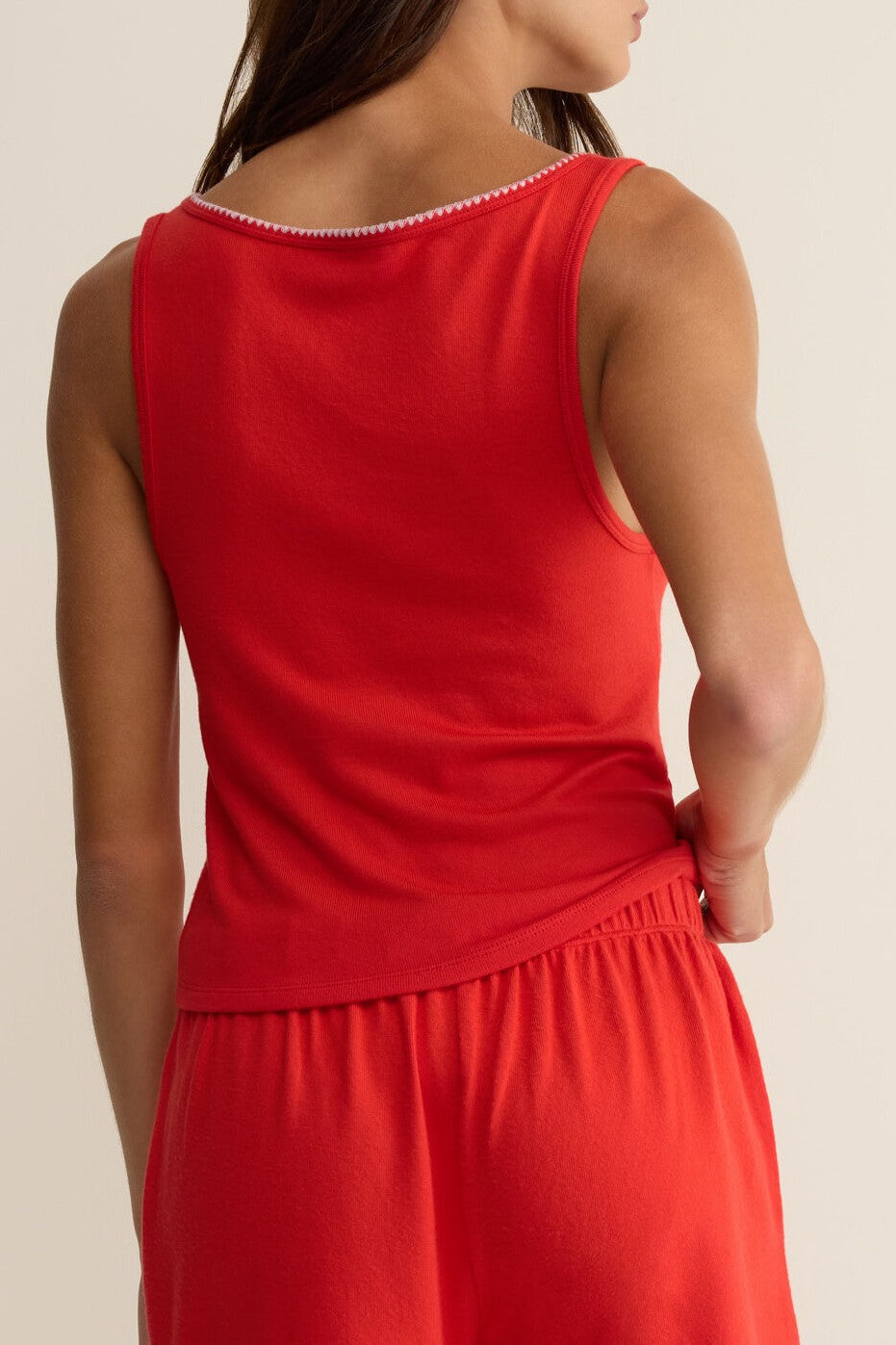 A woman with medium-length brown hair stands with her back to the camera, wearing the Stately V-neck Tank by Z Supply in red, paired with matching shorts. Her left hand rests on her hip against a plain light background.