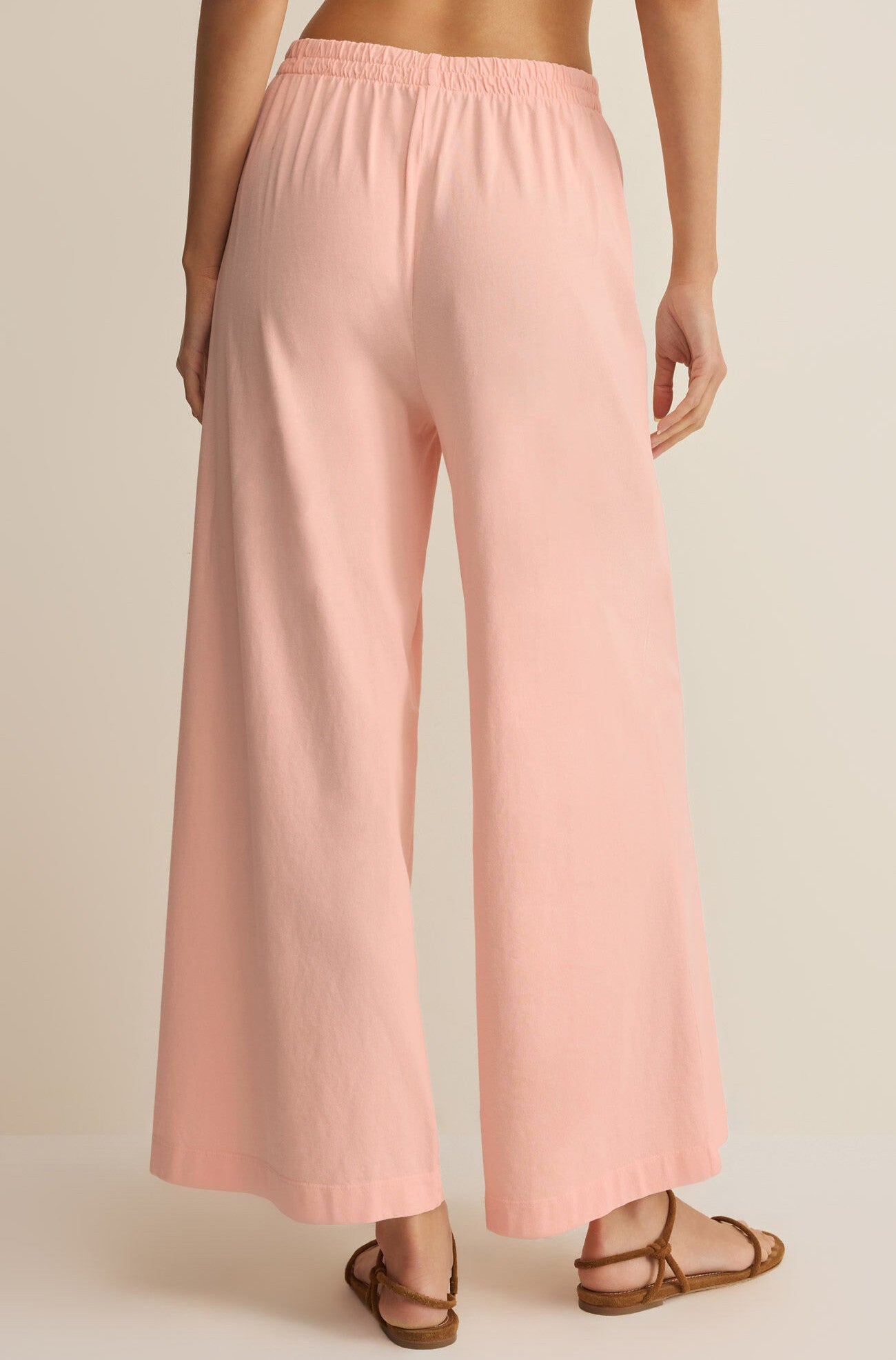 A person faces away, wearing Z Supply's Scout Jersey Crop Flare Pant in relaxed-fit light pink cotton jersey with a wide flare silhouette and brown sandals, set against a plain neutral background.