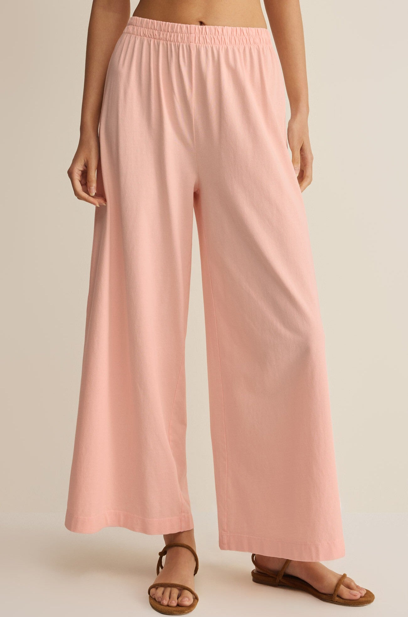 A person wears Z Supply's Scout Jersey Crop Flare Pant in pink, featuring a relaxed fit and elastic waistband, paired with brown flat sandals. The image is cropped from the shoulders down against a plain background.