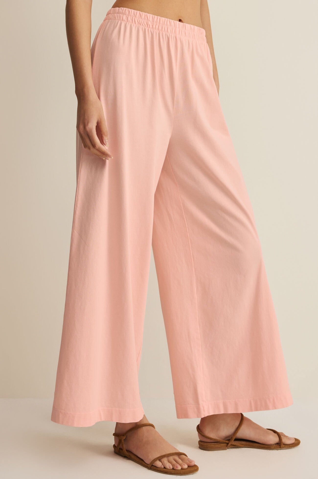 The lower half of a person wearing Z Supply's Scout Jersey Crop Flare Pant in light pink with brown sandals is shown against a neutral background.