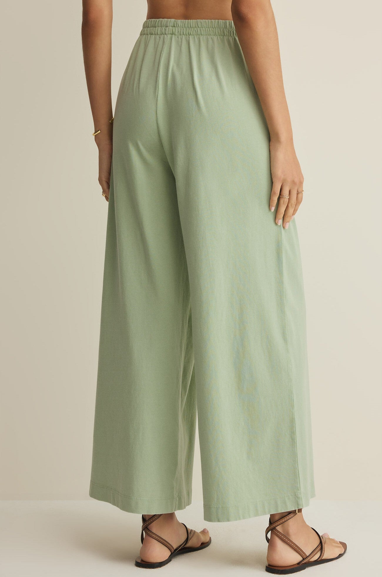 A person stands facing away, wearing Z Supply's Scout Jersey Crop Flare Pant in light green cotton, styled with a gold bracelet, rings, and black strappy sandals against a neutral background.