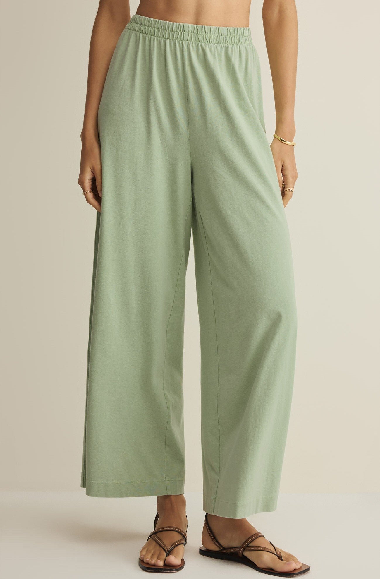 Wearing Z Supply’s Scout Jersey Crop Flare Pant in green, styled with flat black sandals and a gold bracelet, a person stands against a plain light background.