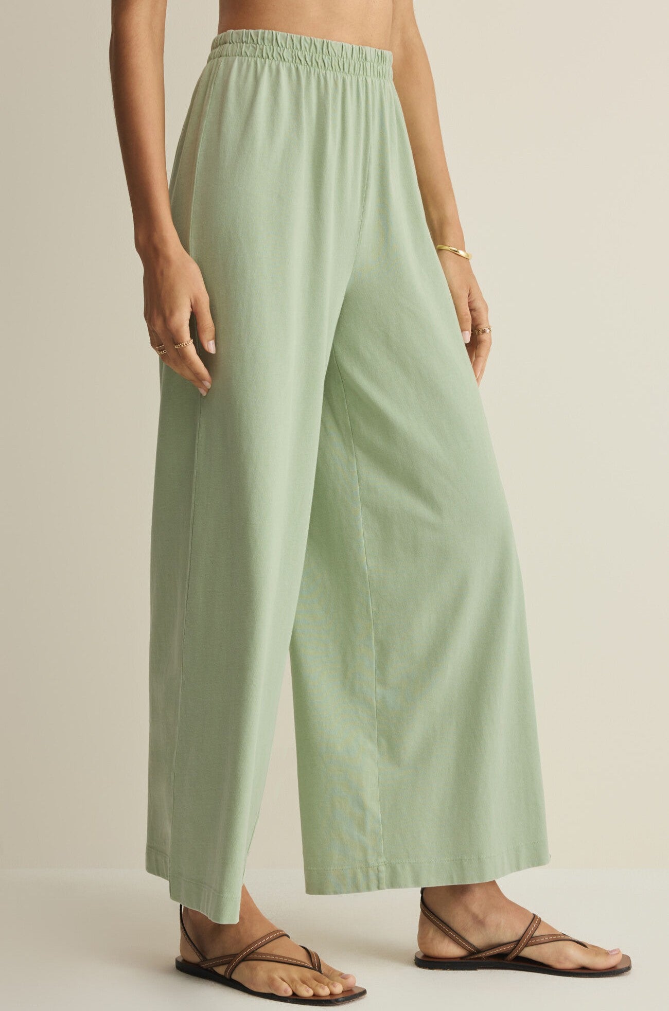 Wearing Z Supply's Scout Jersey Crop Flare Pant in light green cotton with a wide flare and elastic waistband, styled with a gold bracelet and black strappy sandals, the person stands against a plain beige background.