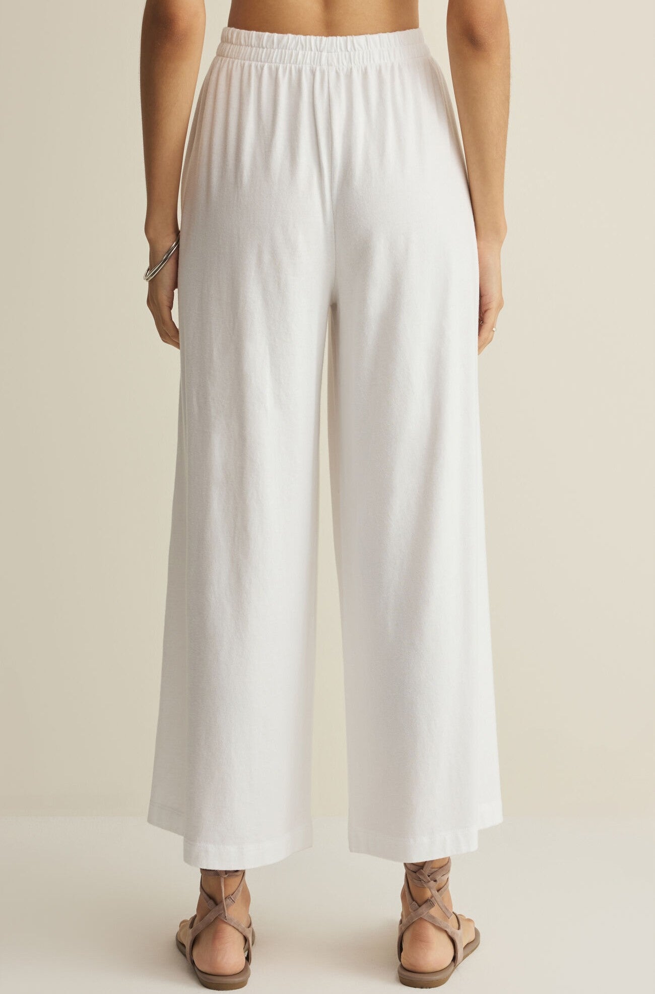 A person stands barefoot, shown from the back, wearing Z Supply’s Scout Jersey Crop Flare Pant—high-waisted with a wide flare silhouette and elastic waistband—paired with a silver bracelet against a neutral background.
