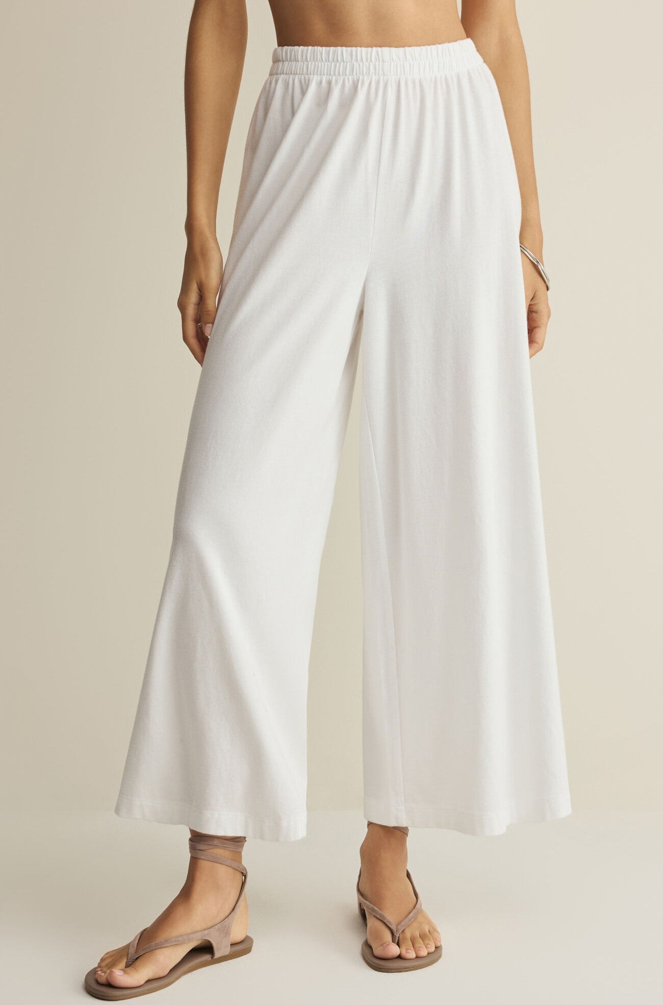 A person models the Z Supply Scout Jersey Crop Flare Pant—a relaxed fit, wide flare white pant with an elastic waistband—paired with tan sandals. Photo from waist down, outfit against a neutral background.