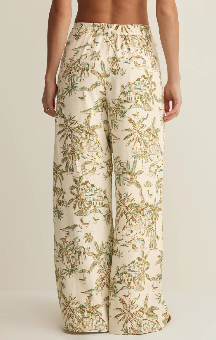 A person is shown from the waist down wearing Z Supply's Overlook Tiki Tide Linen Pant in cream, featuring a high-rise, wide-leg fit and tropical palm tree and house print on washed linen, set against a plain neutral background.