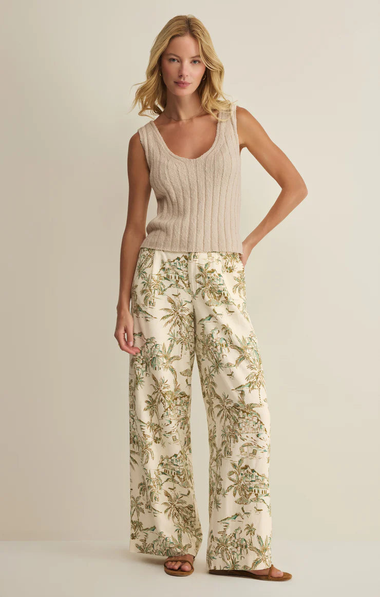 A woman with shoulder-length blonde hair wears Z Supply's Overlook Tiki Tide Linen Pant, featuring a green and brown leafy floral print, paired with a sleeveless beige knit top and brown sandals against a plain background.