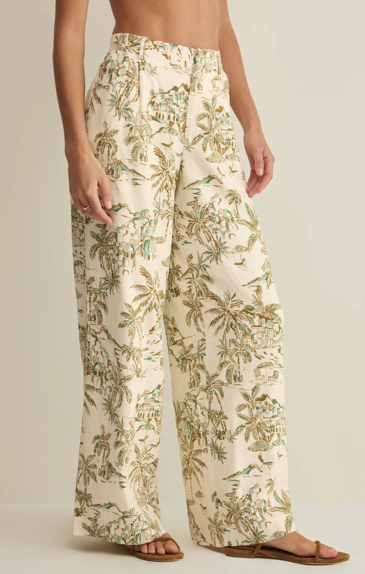 A person wears Z Supply's Overlook Tiki Tide Linen Pant, featuring a cream washed linen with a tropical palm tree and house print, paired with flat sandals. The top half of the person is not visible.