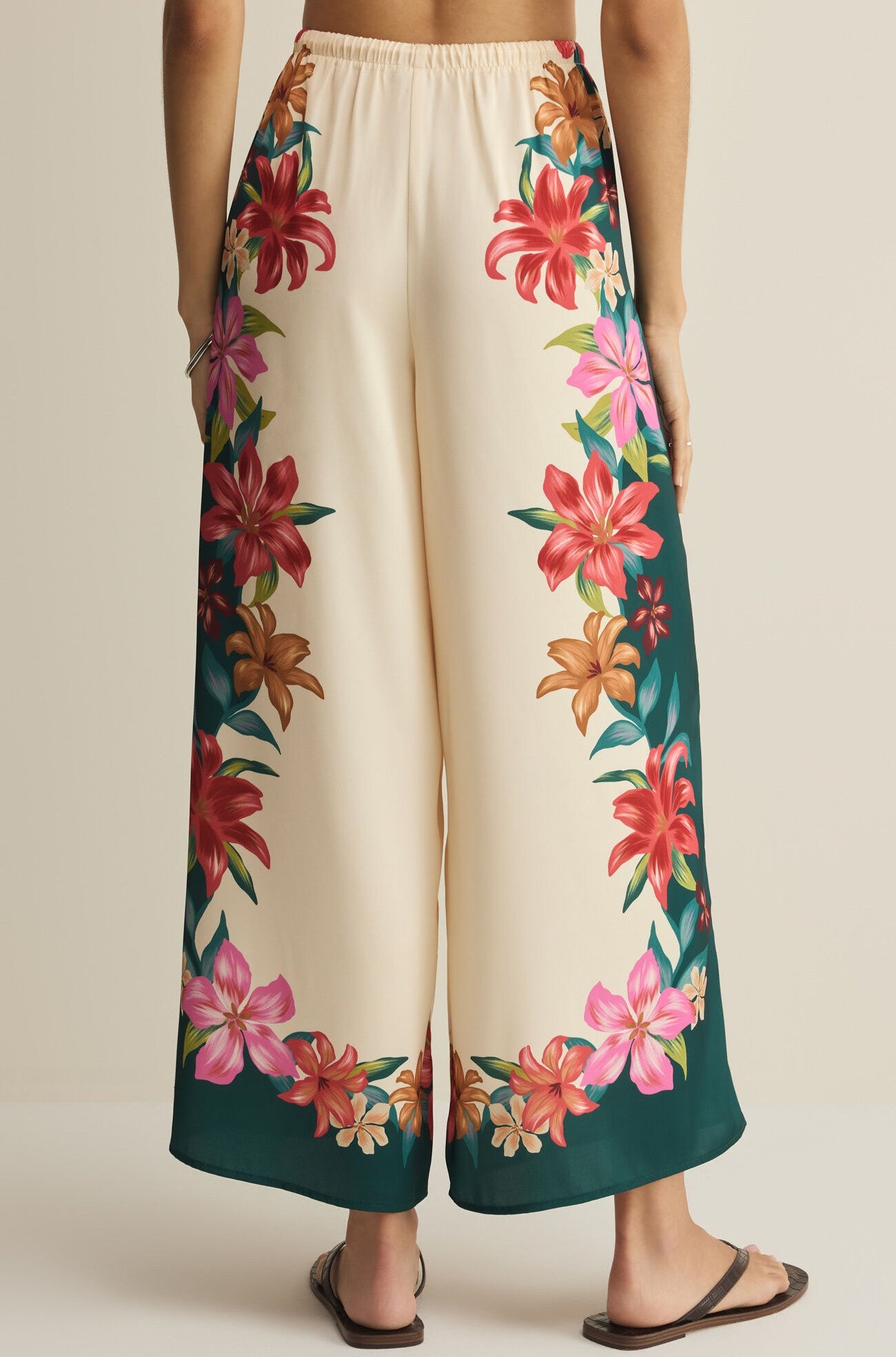 A person seen from the back wears Z Supply's Lani Kiwi Pant—a high rise, wide leg cream pant with a green border and colorful floral prints along the sides and bottom—paired with sandals.