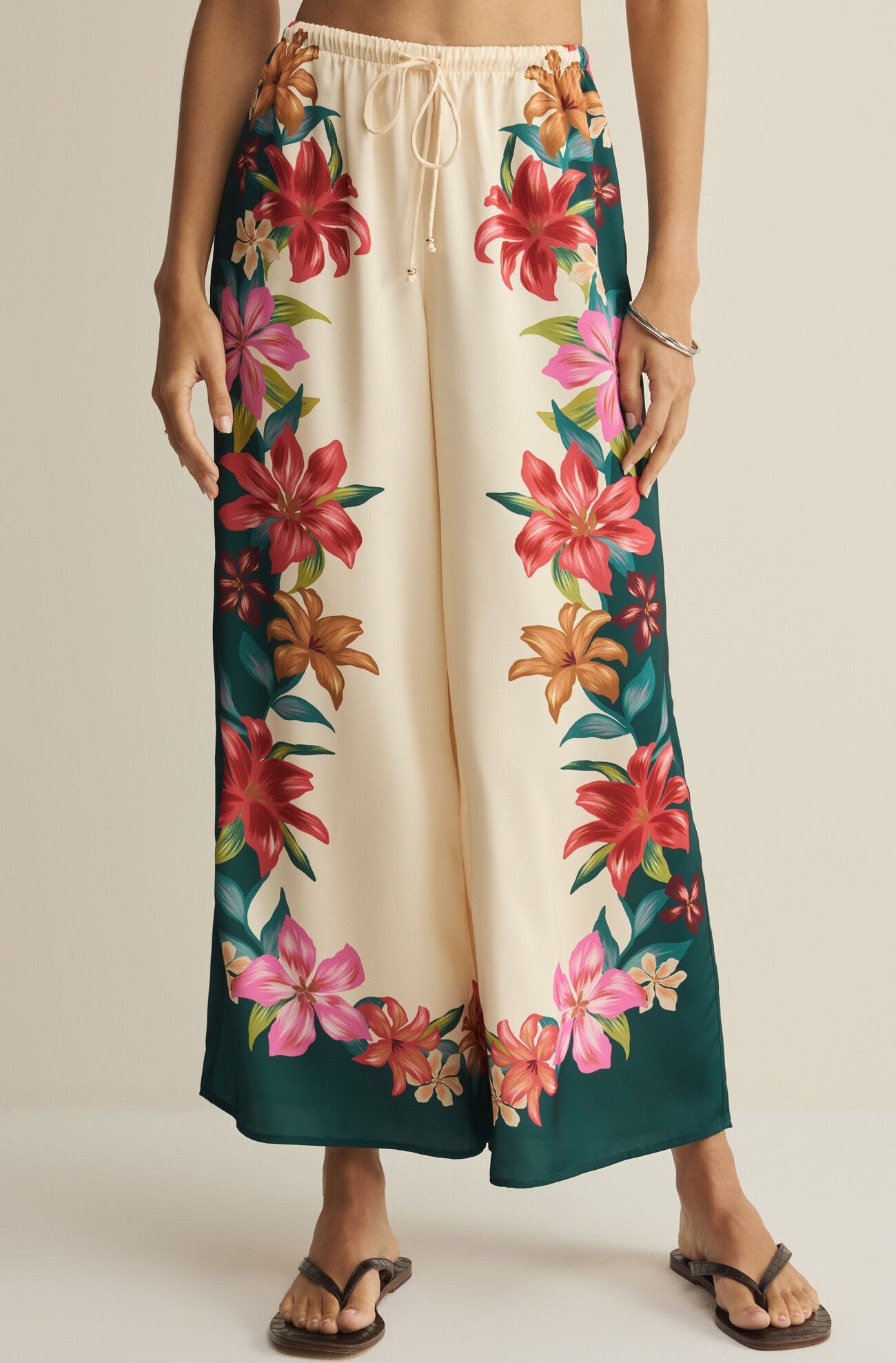 A person wears the Z Supply Lani Kiwi Pant in Multi—a high rise, wide leg linen pant with a drawstring waist, vibrant red, pink, and orange floral prints along the sides, green hem panels—paired with sandals.