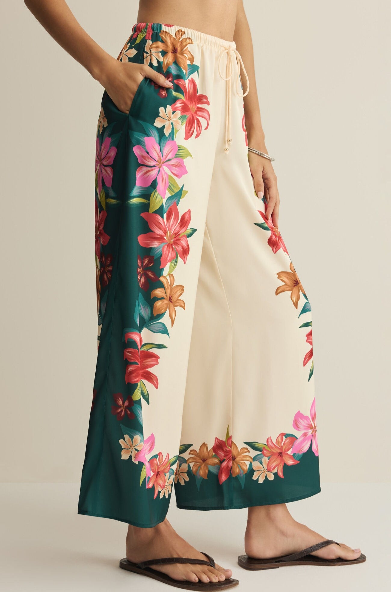 A person wears Z Supply’s Lani Kiwi Pant—high-rise, wide-leg cream pants with a vibrant pink, red, and orange floral border—paired with sandals. Only the lower torso and legs are shown.