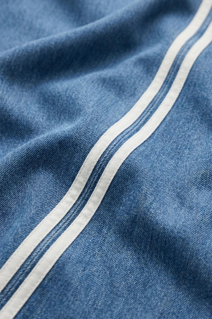 Introducing The Sidelines Denim Track Pant from Z Supply—crafted from 100% cotton blue denim, this pant features two parallel white stripes running diagonally across the fabric. The slightly wrinkled finish highlights its soft texture and relaxed, casual style.