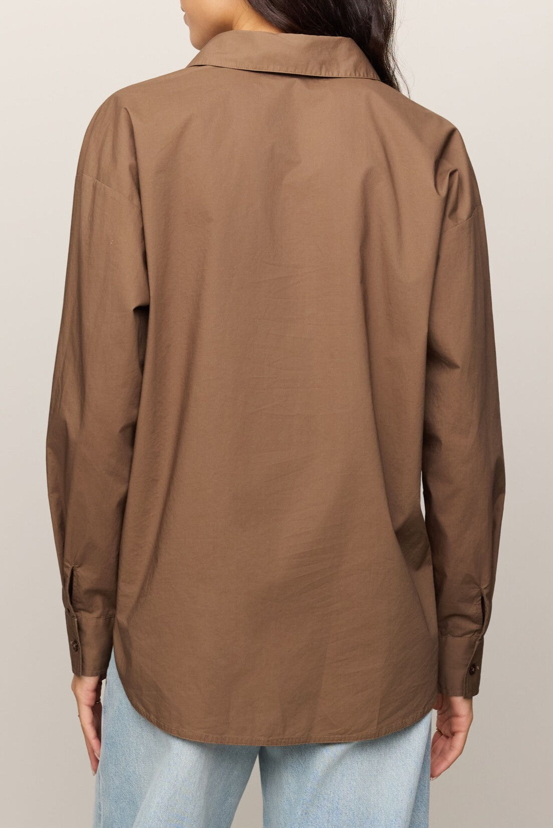 A person is shown from the back against a plain light background, wearing the Z Supply Elevated Poplin Shirt—a collared, long sleeve brown button-up with a relaxed fit—paired with light blue jeans.