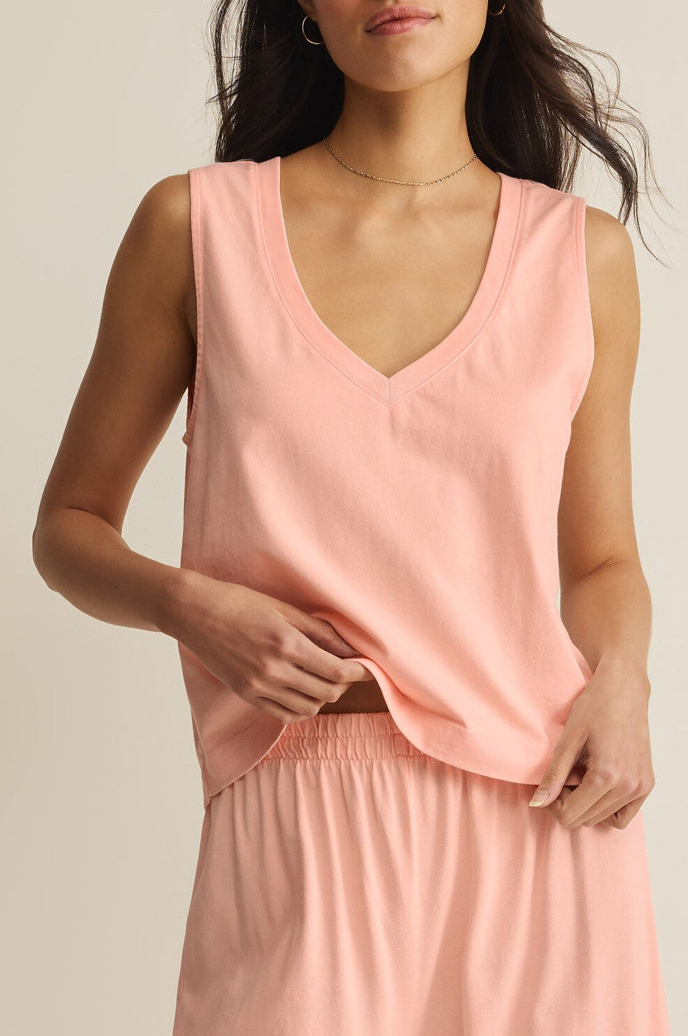 A woman wearing the Z Supply Sloane V-neck Top, a sleeveless light pink relaxed fit tank, with matching elastic-waist shorts stands against a neutral background, partially lifting the hem of her top with one hand.