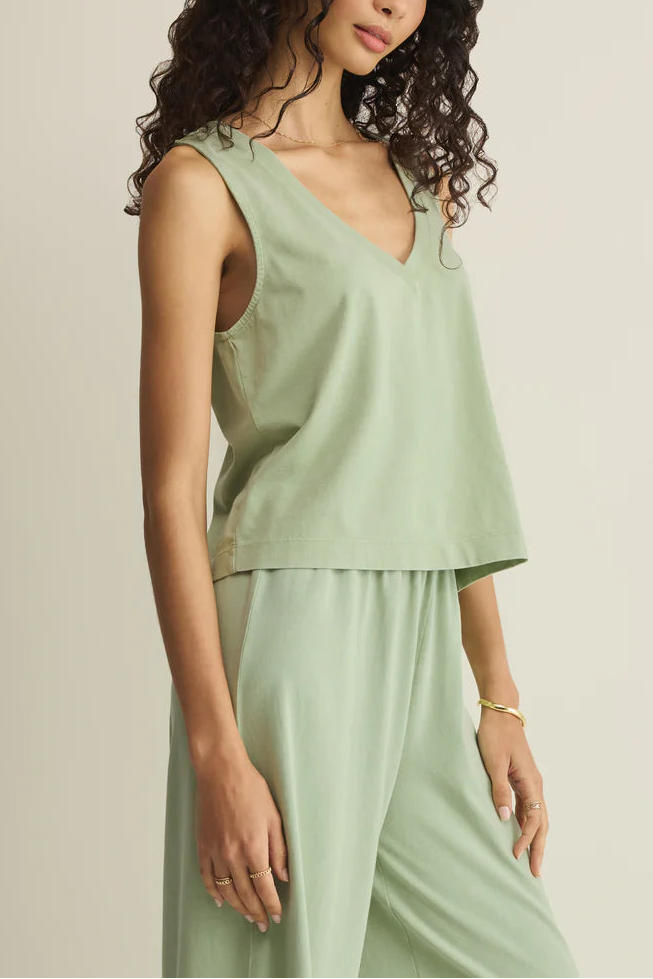 A woman with curly hair wears the Z Supply Sloane V-neck Top in Crystal Green and matching relaxed pants, accessorized with simple gold jewelry. She stands with one arm by her side against a plain, light background.
