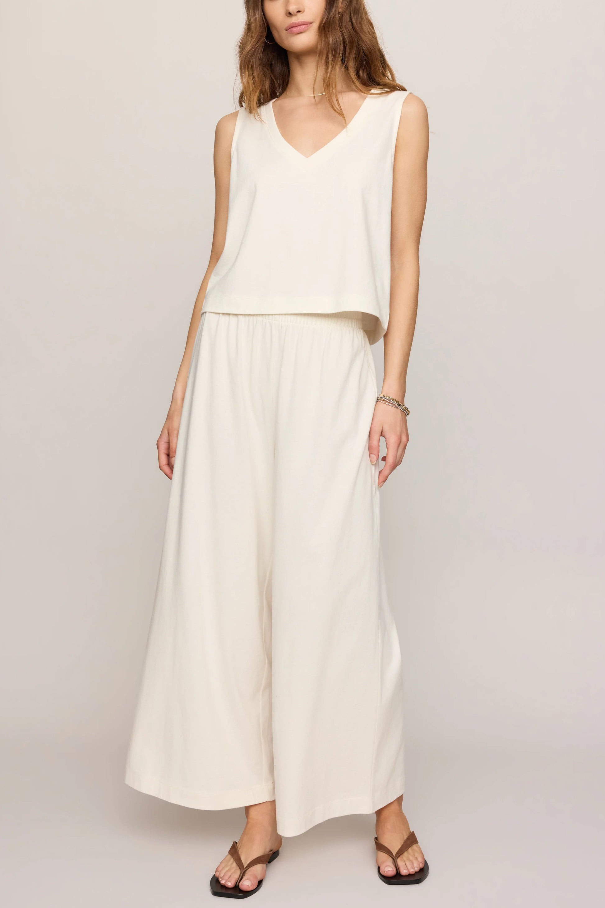 A woman wears the Z Supply Sloane V-neck Top in Sea Salt and matching wide-leg pants, standing against a plain light background. She has wavy hair and accessorizes with sandals and minimal jewelry.