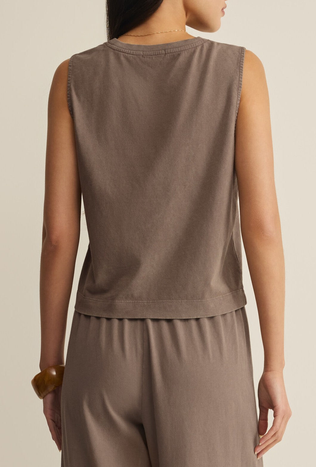 A person is seen from the back wearing the Z Supply Sloane Jersey Muscle Tank in taupe with matching relaxed-fit pants and a brown bracelet. The background is plain and neutral.