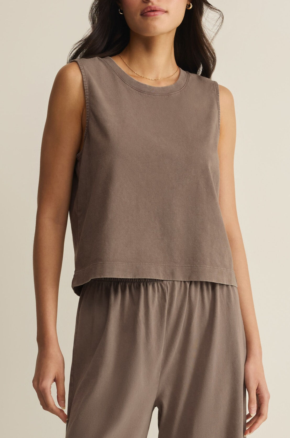 A woman wears the Sloane Jersey Muscle Tank by Z Supply in brown with matching high-waisted pants, standing against a neutral background. She accessorizes with small hoop earrings and a thin necklace, and her dark hair is partially visible.