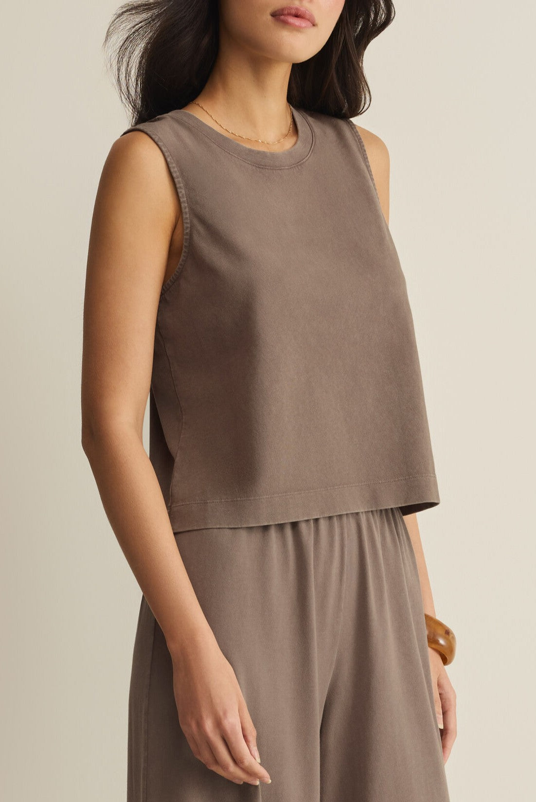 A woman wears the Z Supply Sloane Jersey Muscle Tank, a sleeveless, relaxed-fit taupe cotton crop top, with matching wide-leg pants, standing with one hand by her side against a plain background.