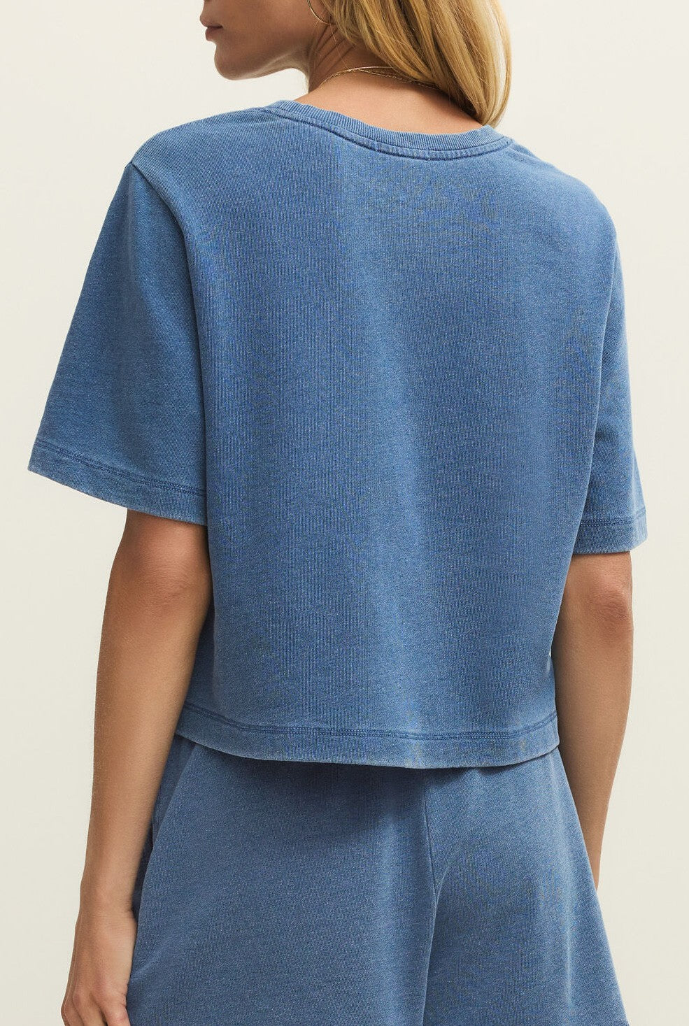 A woman with blonde hair, seen from the back, wears the Z Supply Curran Knit Denim Sweatshirt and matching blue shorts made of knit denim French terry. The relaxed fit outfit stands out against a plain, light-colored background.