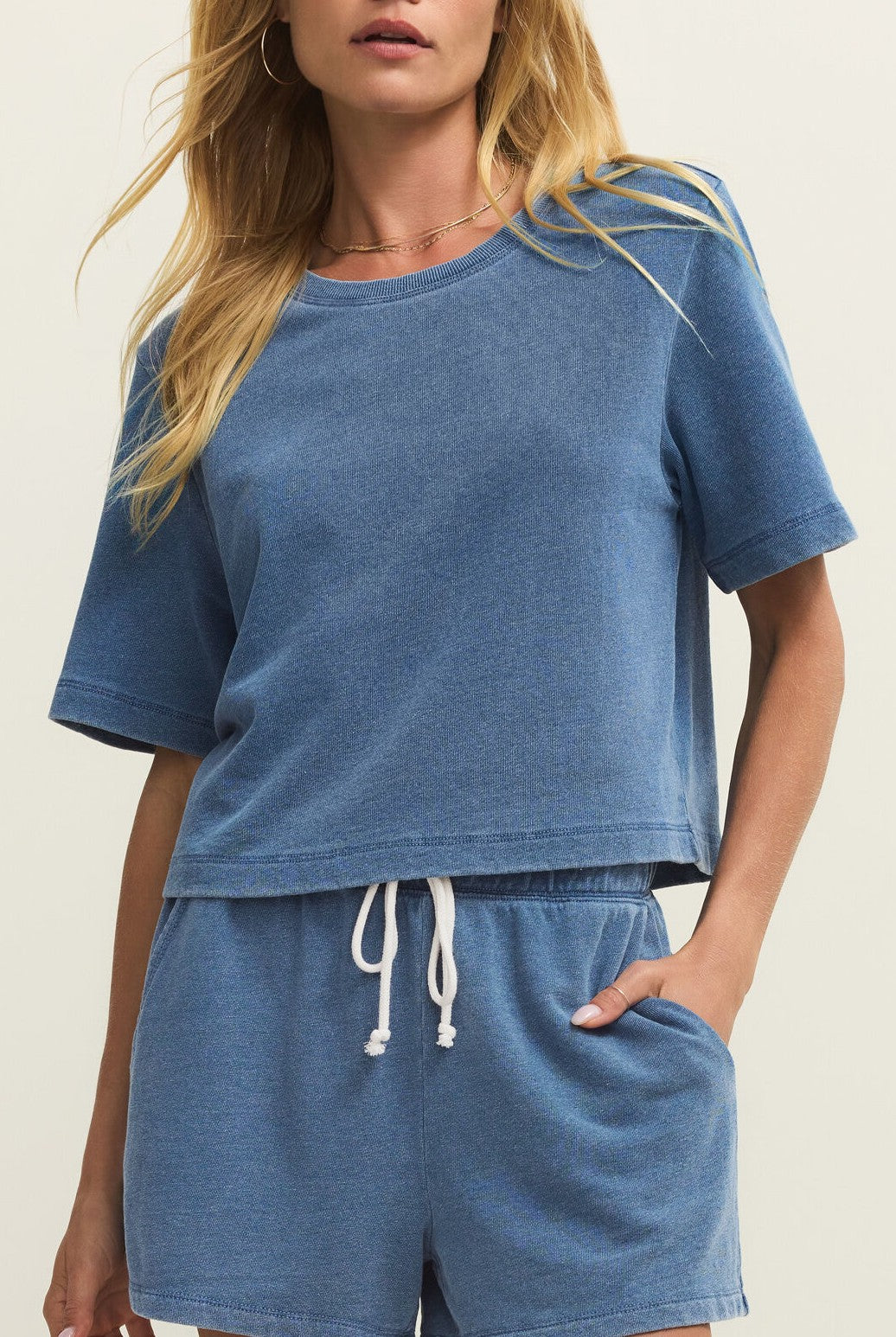 A woman wears the Z Supply Curran Knit Denim Sweatshirt in blue, styled with matching drawstring shorts. With her right hand in her pocket, the soft set looks relaxed and comfortable.