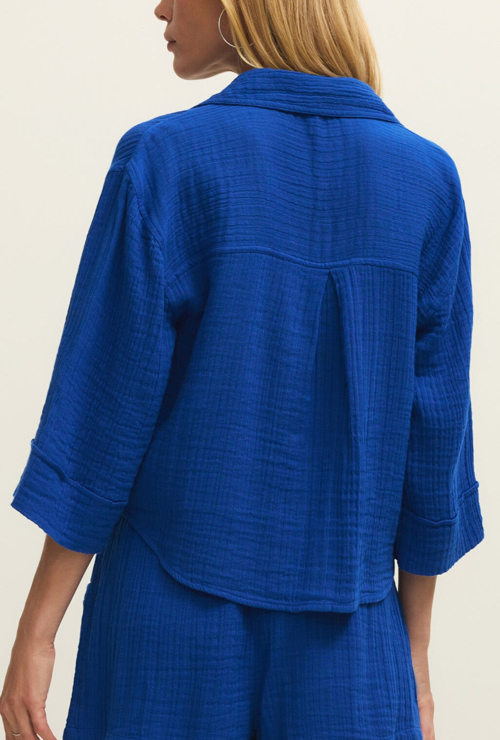 A woman with blonde hair is seen from the back wearing the Z Supply Helena Gauze Shirt in bright blue cotton with three-quarter sleeves and matching shorts, set against a plain light background.