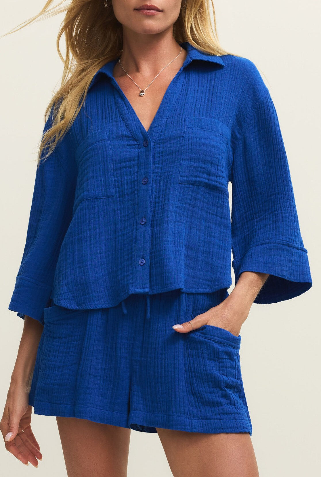 A woman wears the Z Supply Helena Gauze Shirt in royal blue—a textured cotton top with a button front and front pockets—styled as a matching set, standing against a plain light background.
