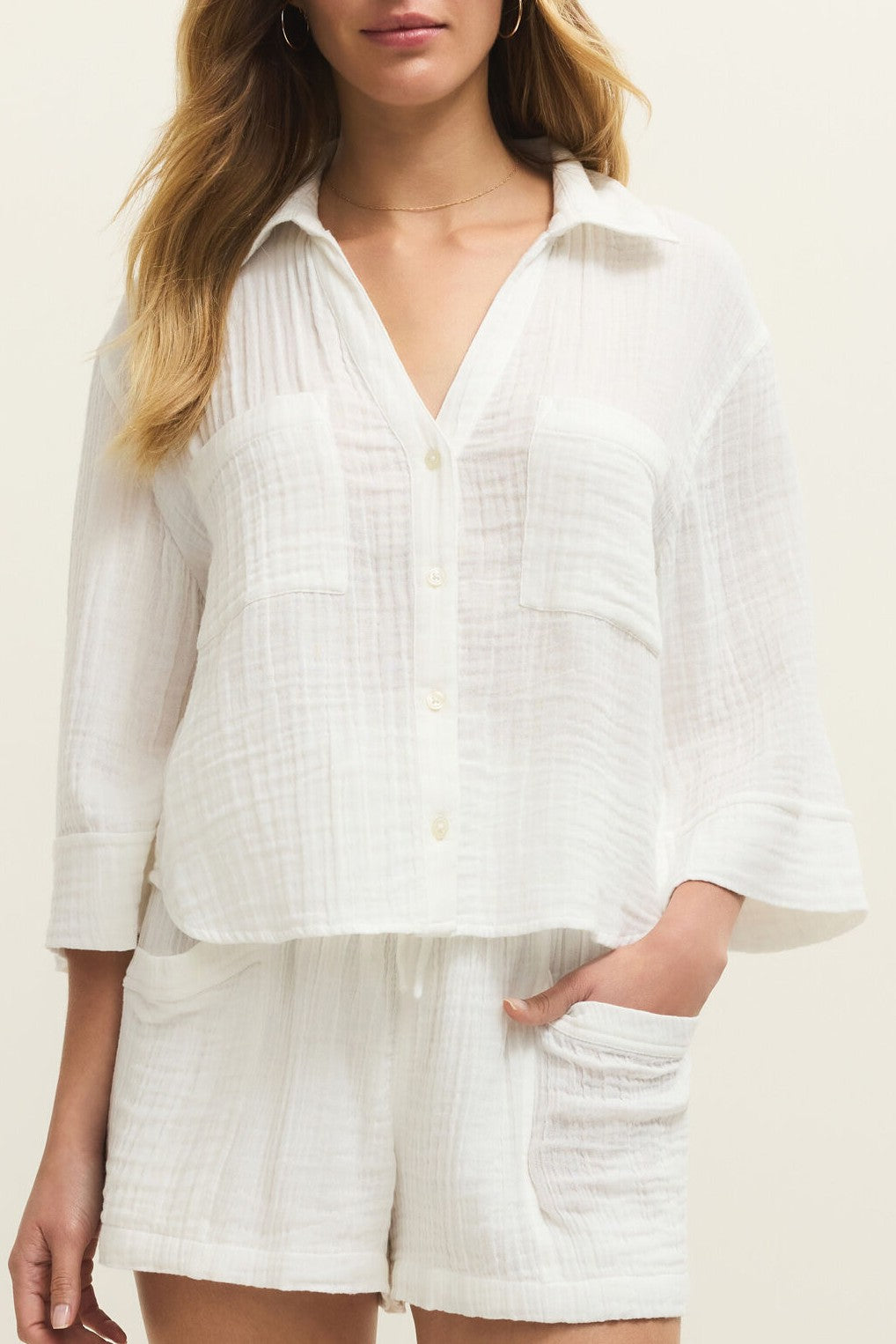 A woman wears the Z Supply Helena Gauze Shirt, a textured, loose-fit white cotton button-front top with long sleeves, paired with matching white shorts. She stands against a plain background with one hand in her shorts pocket.