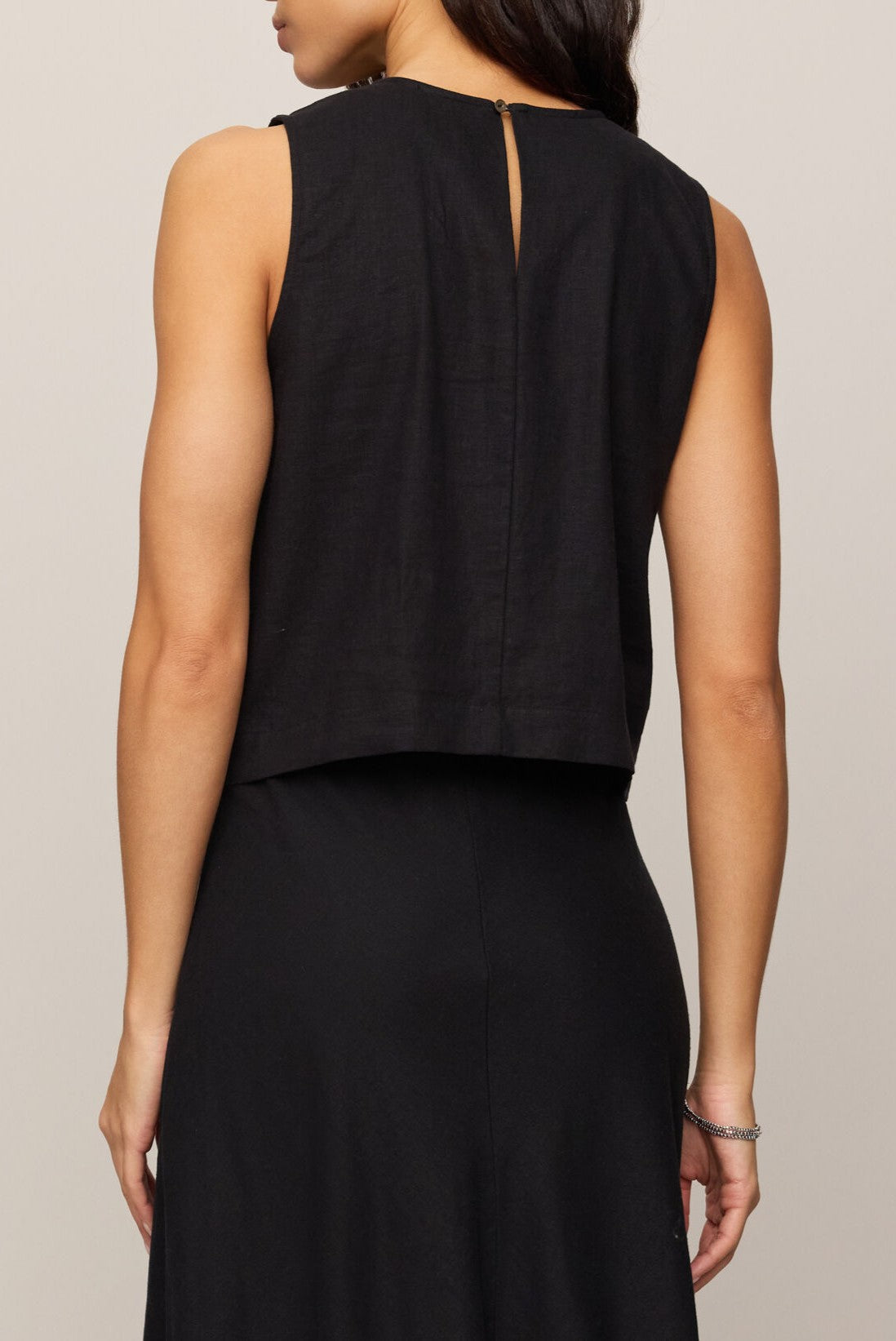 A woman with long, wavy hair and a silver bracelet on her right wrist wears the Z Supply Sloane Linen Top—a sleeveless black linen blend tank with a keyhole button closure—paired with a matching black skirt, highlighting its relaxed fit.