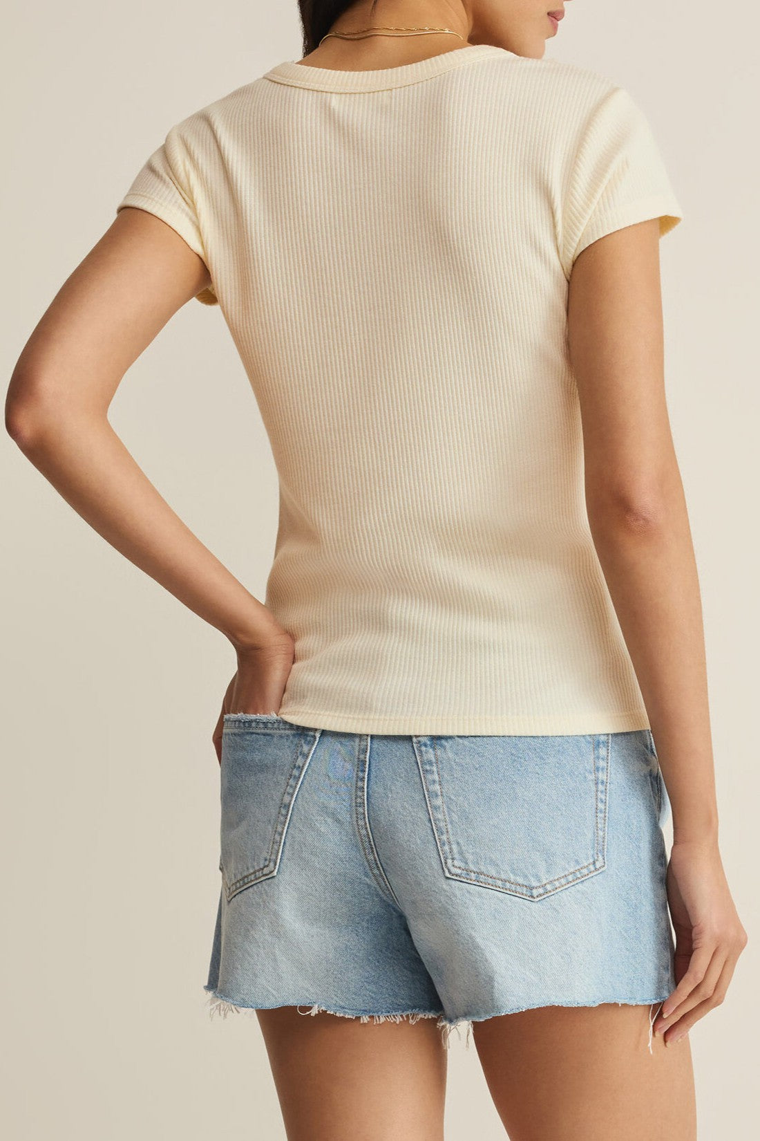 Someone wearing the Z Supply Leisurely Tee, a short sleeve ribbed cream top with a regular fit, and light blue denim shorts stands with one hand on their hip, facing away from the camera.