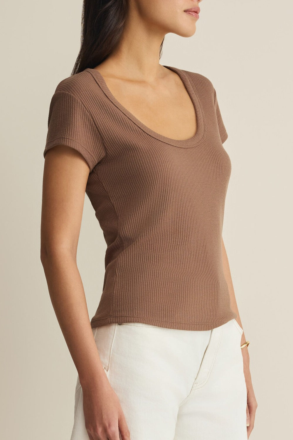 A woman stands sideways with her head turned, wearing the Z Supply Leisurely Tee—a regular fit, short sleeve brown scoop neck top—paired with white pants against a neutral background.
