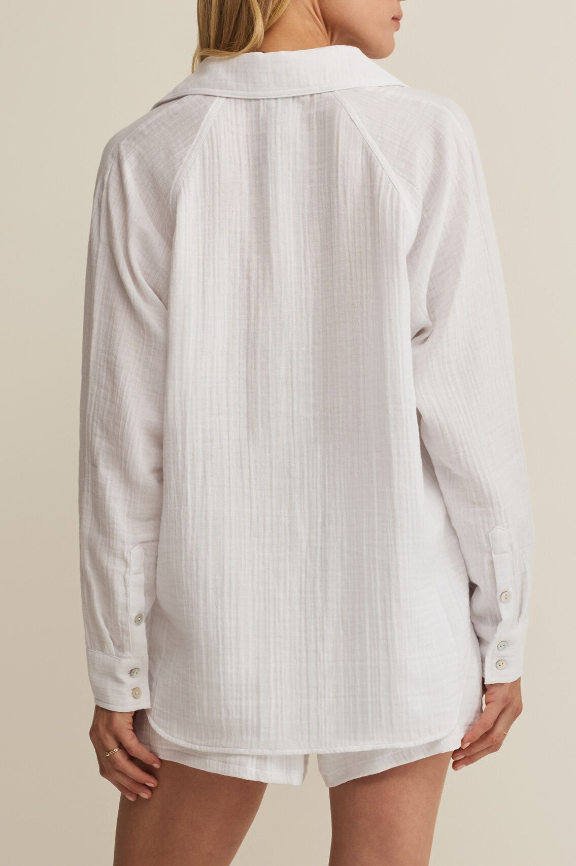 A woman is seen from the back in The Perfect Airy Top by Z Supply, a regular fit, collared long sleeve white button-up shirt, paired with matching white shorts against a neutral background.
