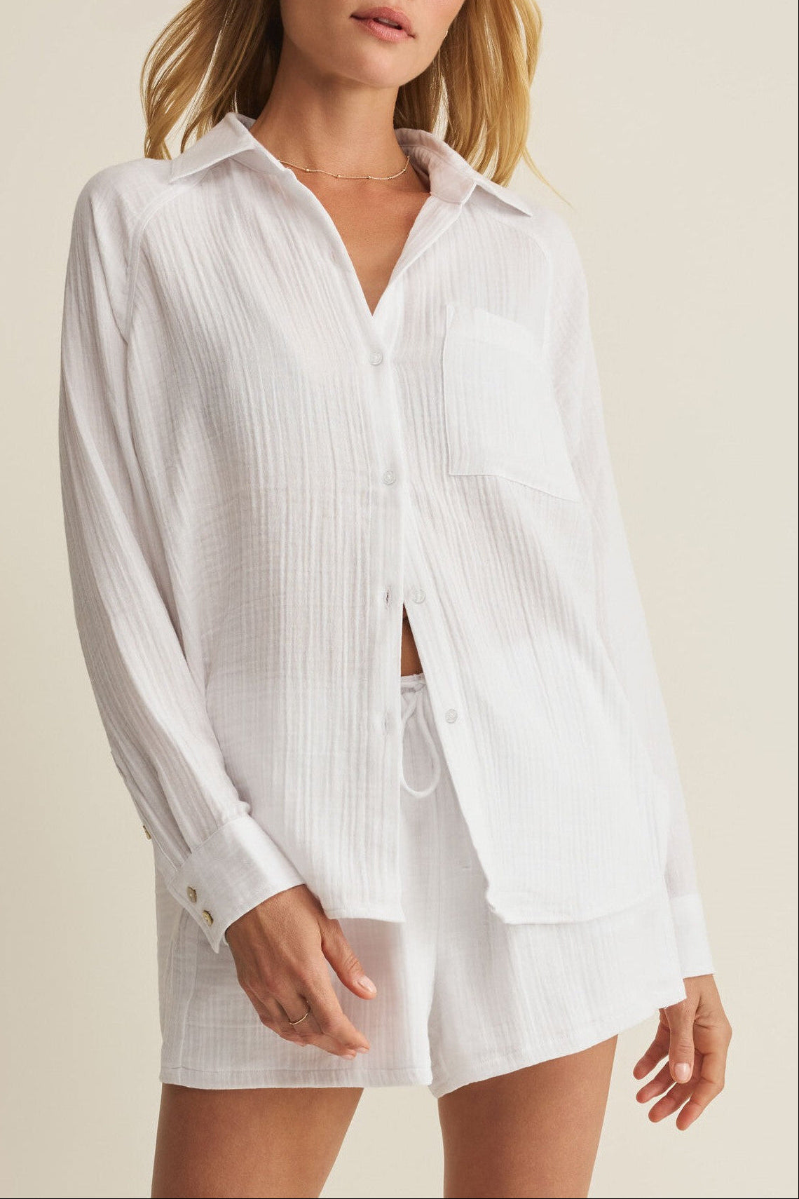A woman wears Z Supply’s The Perfect Airy Top in WHITE—a regular fit, collared long sleeve button front shirt with a breast pocket, partially unbuttoned—paired with matching white shorts against a neutral background.