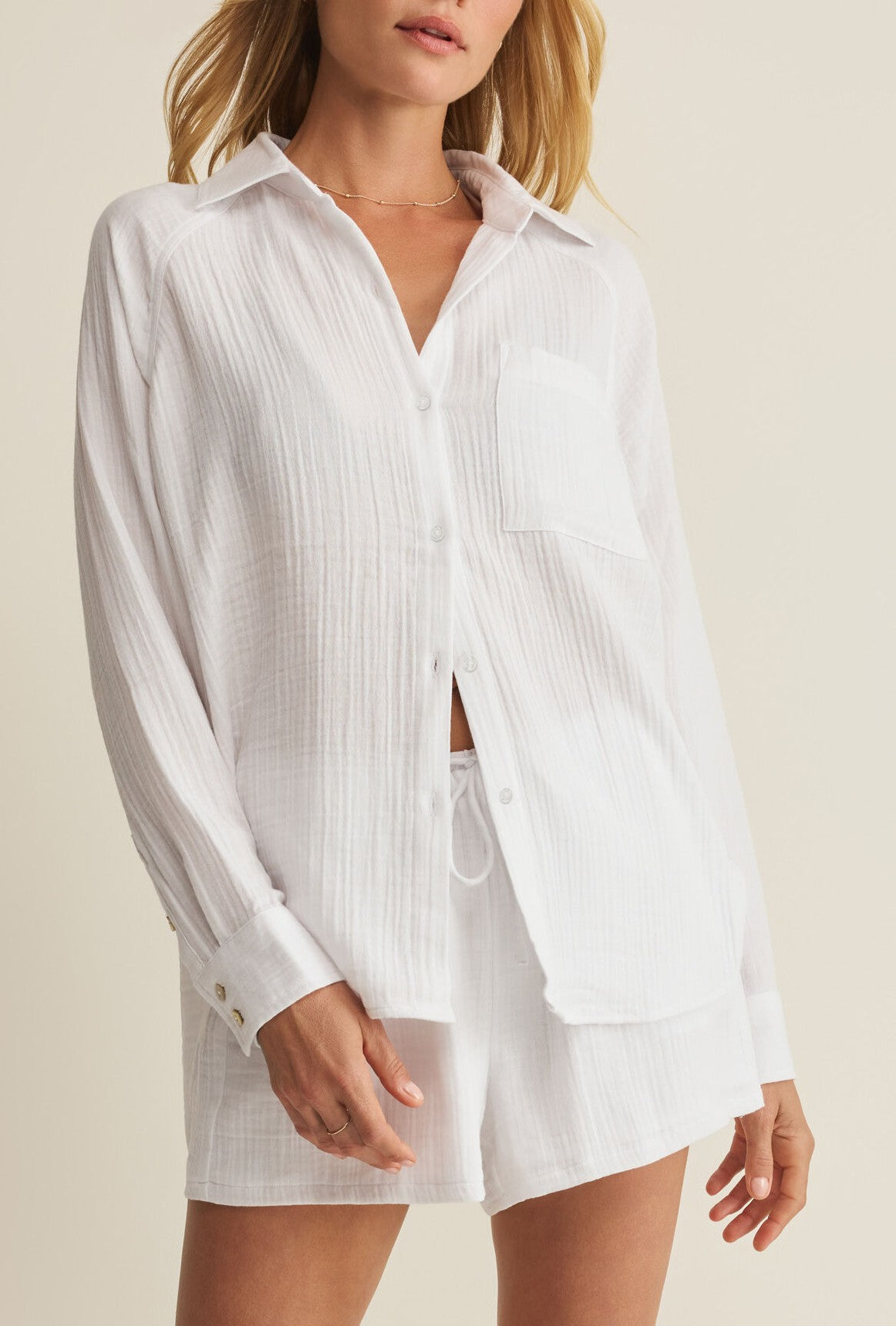A woman wears Z Supply’s The Perfect Airy Top in WHITE—a regular fit, collared long sleeve button front shirt with a breast pocket, partially unbuttoned—paired with matching white shorts against a neutral background.