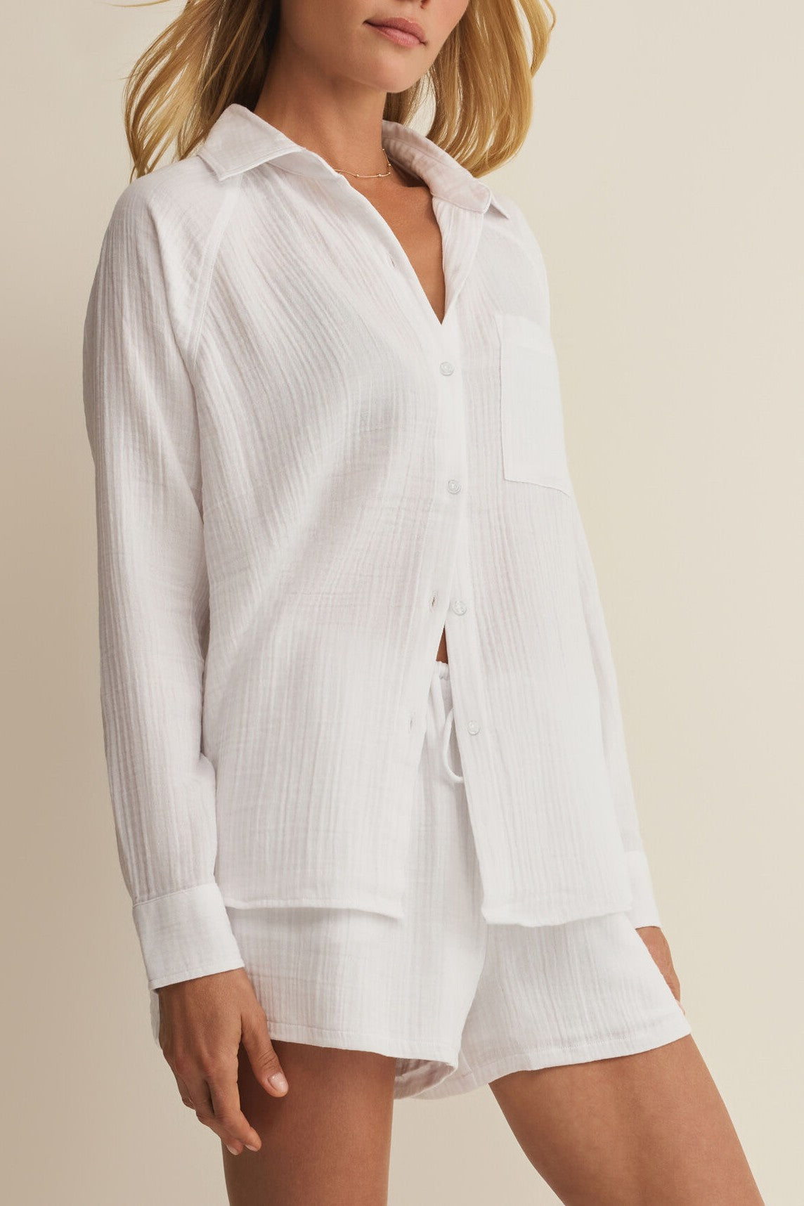 A woman wears Z Supply’s The Perfect Airy Top, a relaxed, collared, long sleeve white button-up with a chest pocket, paired with matching drawstring shorts. This regular fit set is lightweight and comfortable—ideal for lounging or sleepwear.