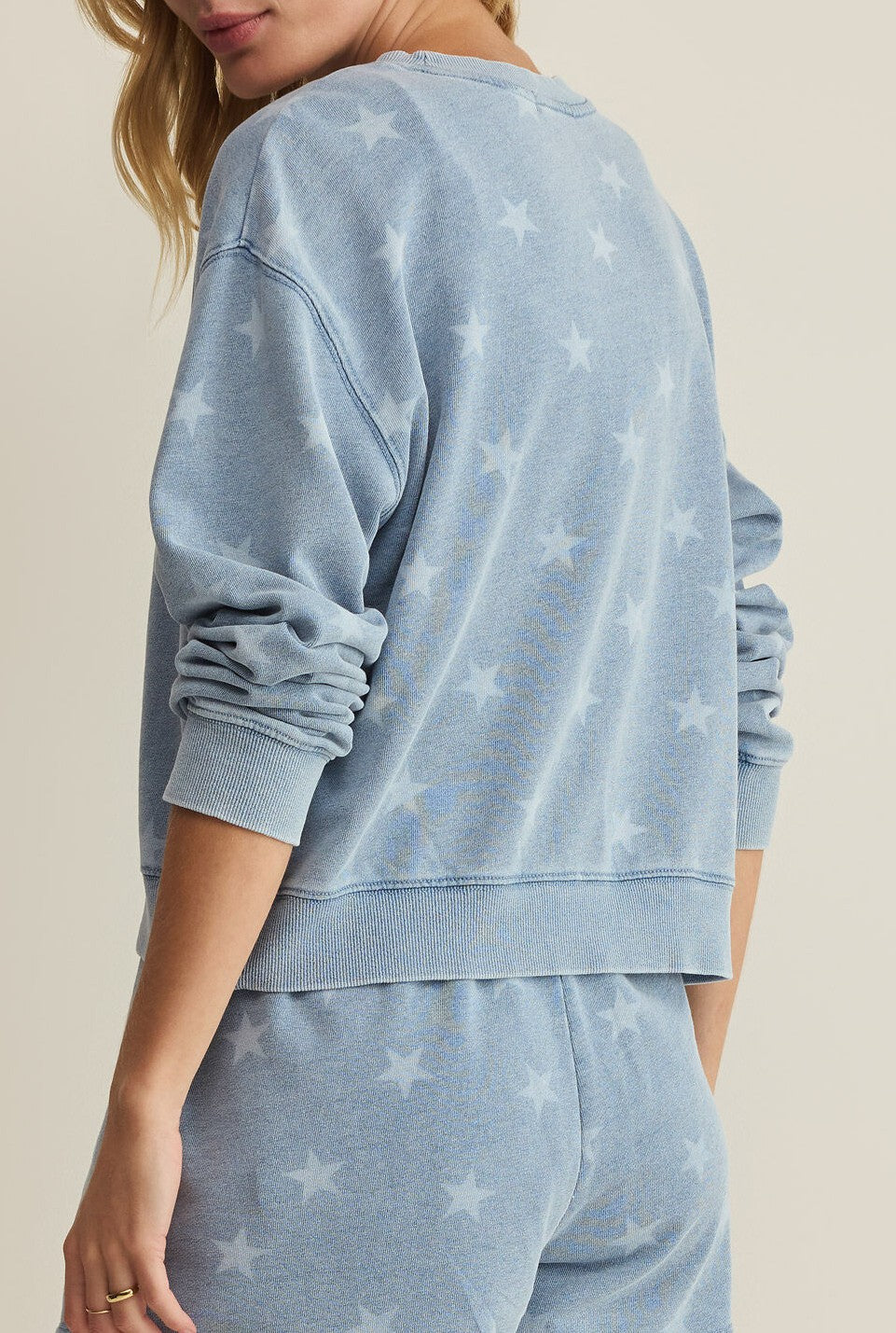 A person is shown from the back and side wearing Z Supply’s Written Stars Sweatshirt—a light blue, long-sleeve crew neck with a subtle star pattern. The relaxed-fit lounge set features partially visible face and comfortable pants.