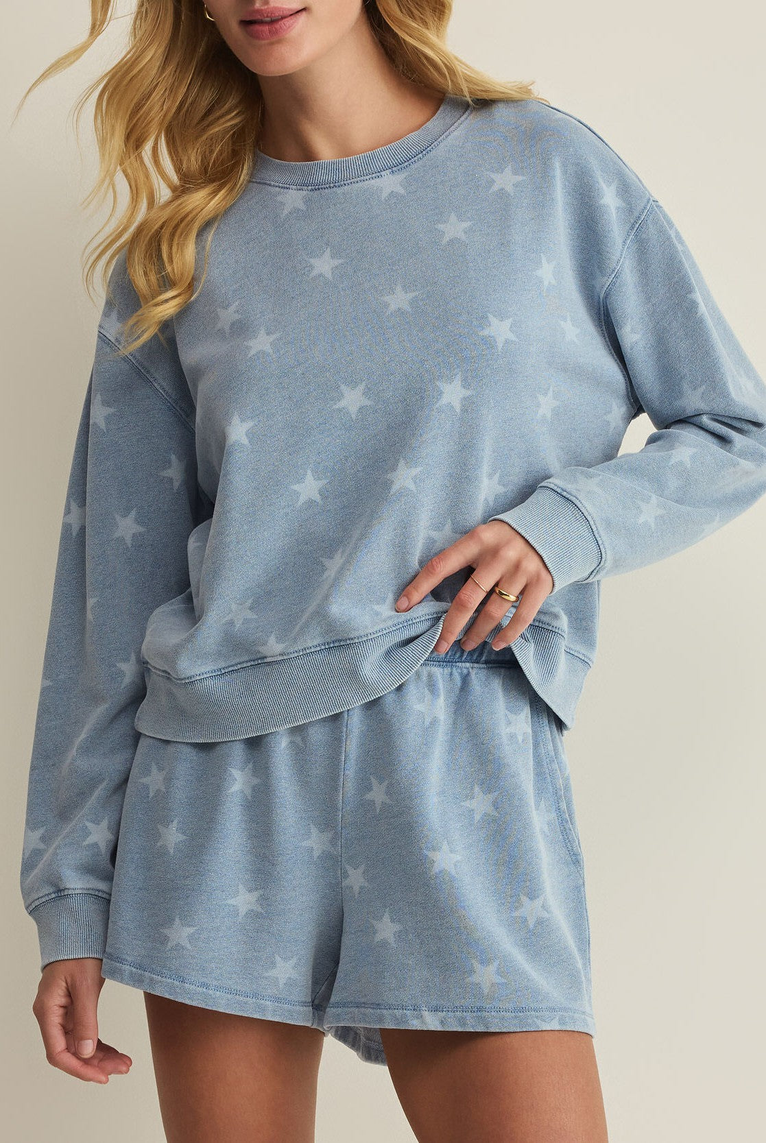 A woman with long blonde hair wears the Z Supply Written Stars Sweatshirt in Blue Multi— a light blue crew neck with a white star pattern—paired with matching shorts, standing against a plain background.