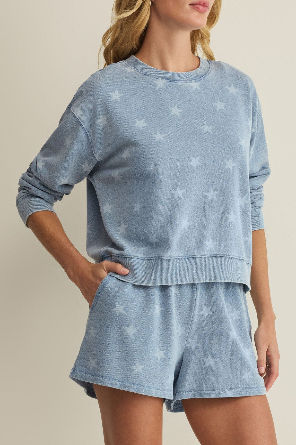 A woman models the Z Supply Written Stars Sweatshirt, a light blue crew neck featuring a subtle star pattern, paired with matching shorts as she poses with one hand in her pocket against a neutral background.
