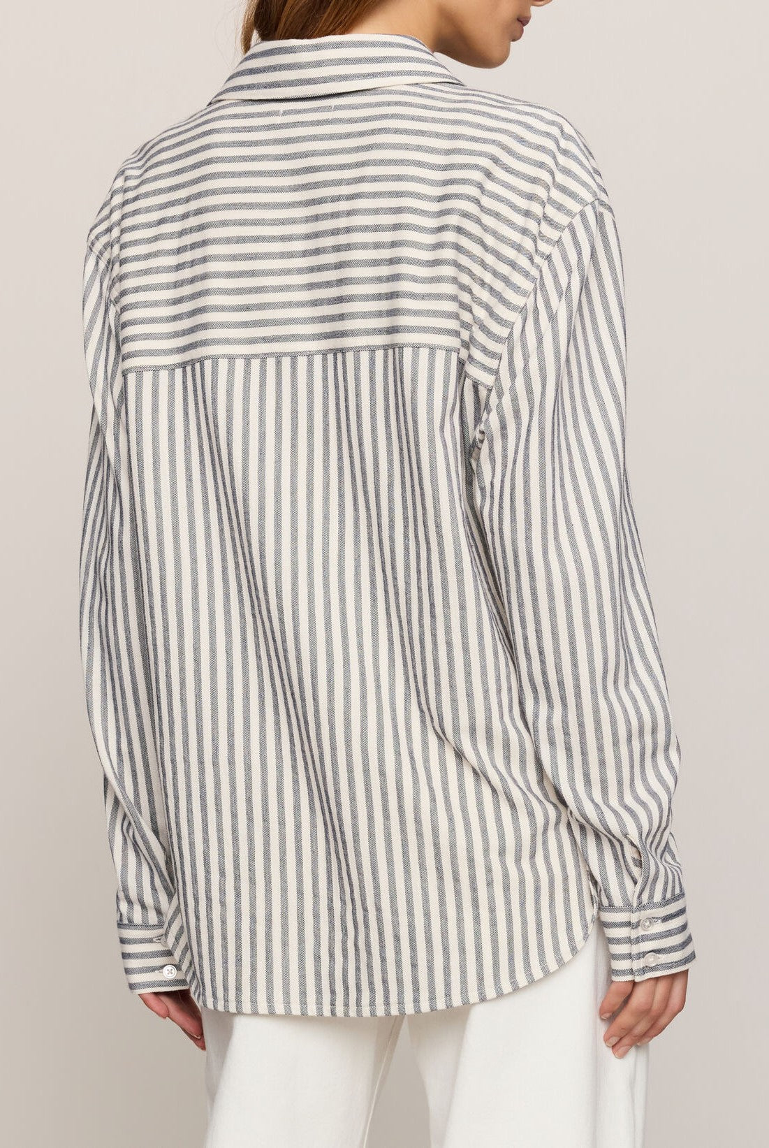 A person with light brown hair pulled back is shown from the back wearing Z Supply’s Reese Button Up Stripe Top—a relaxed fit, long-sleeve white shirt with vertical gray stripes—paired with white pants.