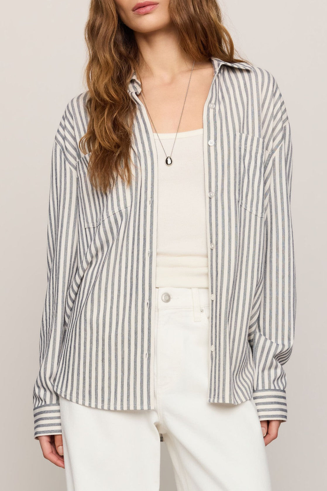 A woman wears the Z Supply Reese Button Up Stripe Top in Black Multi over a white tank and high-waisted white pants. Her wavy hair and simple necklace complete her effortlessly chic style.